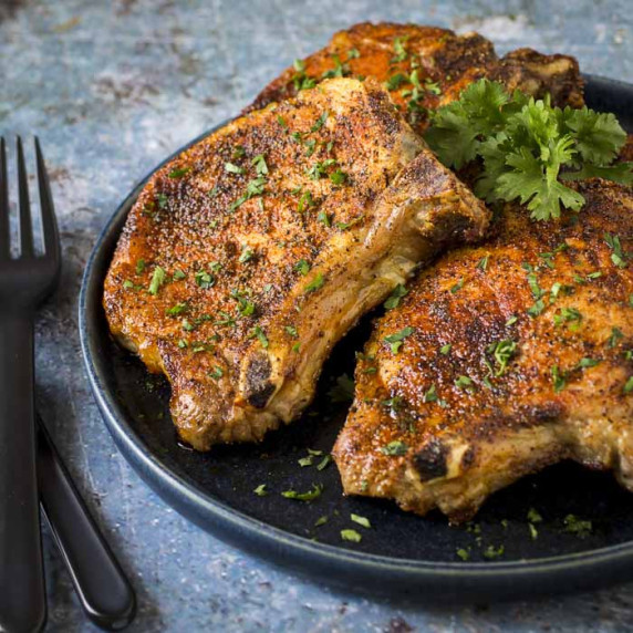 Side shot of three pork chops on a black plate garnished with fresh parsley with a black fork.