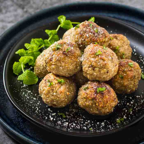 Side shot of multiple meatballs stacked on a black plate garnished with herbs and parmesan cheese.
