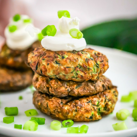 Side shot of three zucchini fritters stacked on each other with a dollop of sour cream and green oni