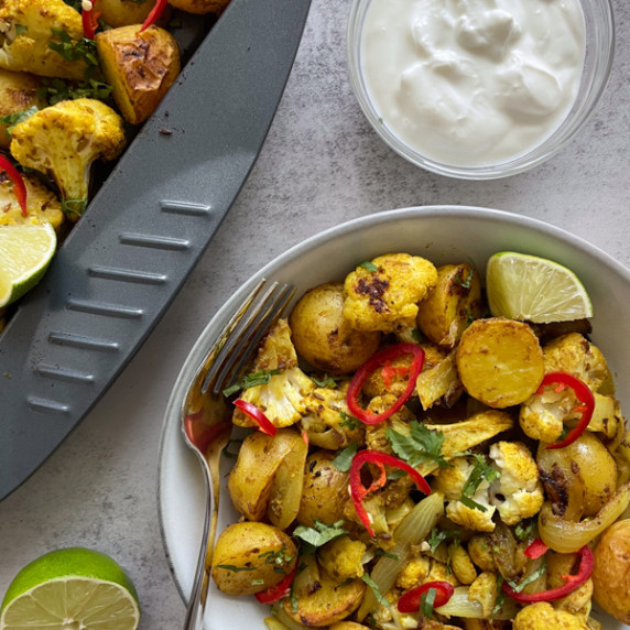 Aloo gobi in a bowl with red chillis, lime and a fork next to a grey tray and bowl of yogurt.
