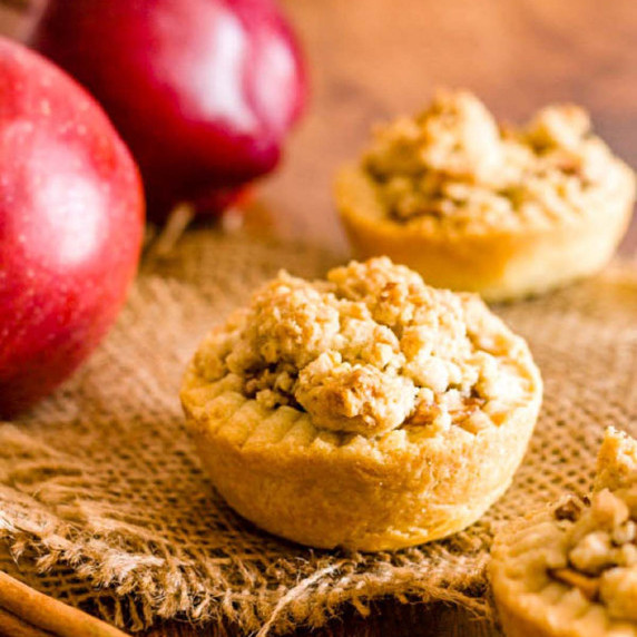 Apple pie tartlets on burlap with apples and cinnamon sticks in the background.