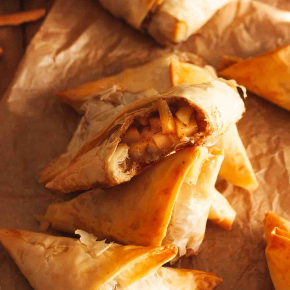 A bitten apple turnover with phyllo dough on top of other turnovers on brown parchment paper.