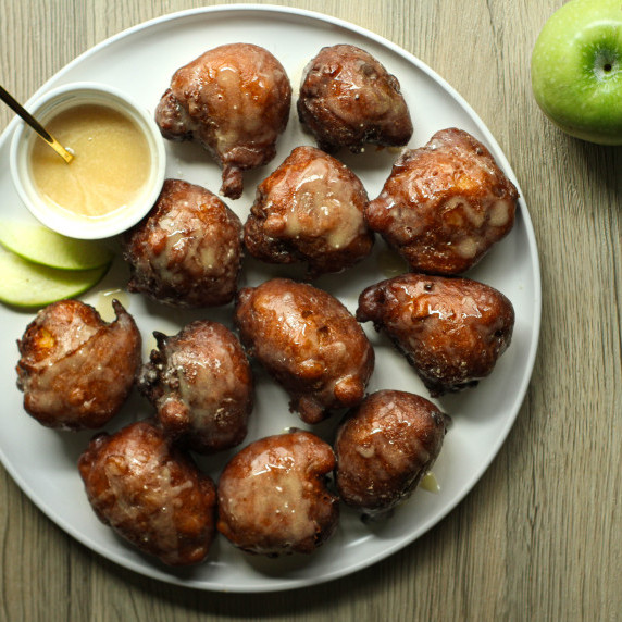 Applesauce fritters drizzled with glaze on a white plate with an apple in the photo.