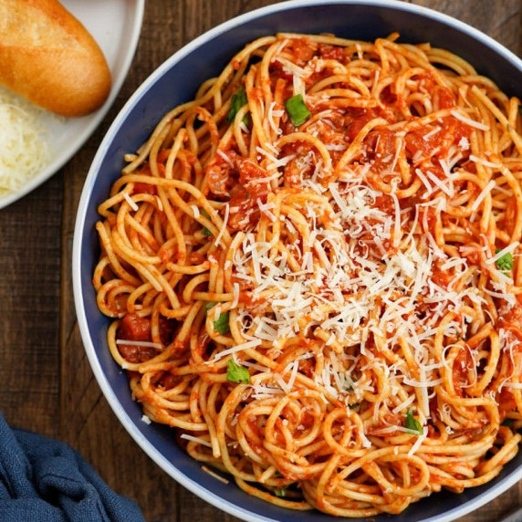 A bowl of spicy pasta arrabiata topped with parmesan cheese on a wooden table with bread along side.
