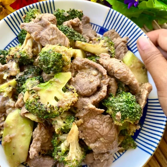 Hand presenting a white dish with stir-fried Thai beef and broccoli.