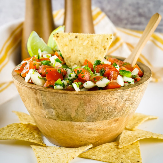 Side shot of a tortilla chip, lime wedges, and a wooden spoon sticking out of a bowl of pico.