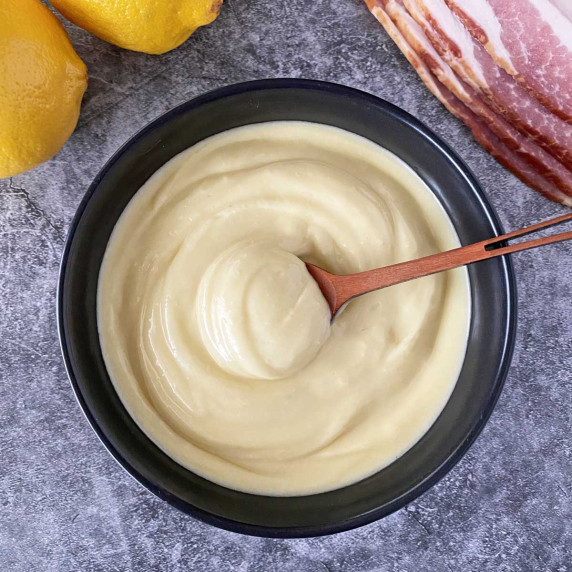 A black bowl of bacon aioli sauce with two lemons and sliced bacon.