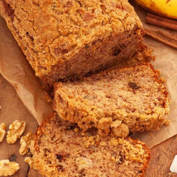 Sliced banana bread with streusel topping on parchment paper next to bananas and walnuts.