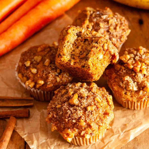 A bitten banana and carrot muffin on top of four muffins, next to carrots and bananas.