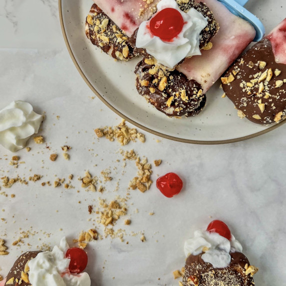 Banana split frozen yogurt pops with whipped cream and cherries on top on a plate with one held up