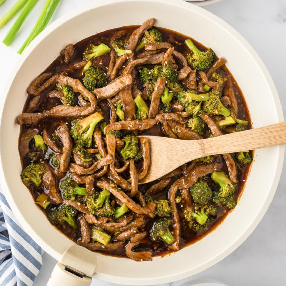 A skillet filled with stir-fried beef and broccoli in a dark sauce with a wooden spoon stirring.