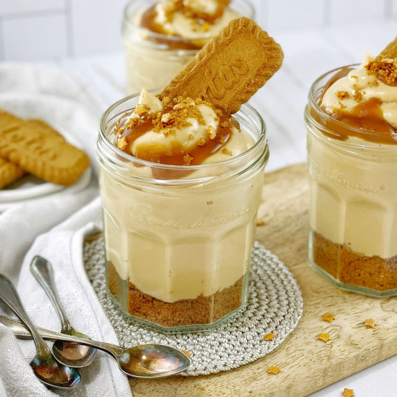 biscoff cheesecake in a clear glass jar with a cookie on top on wooden board with spoons