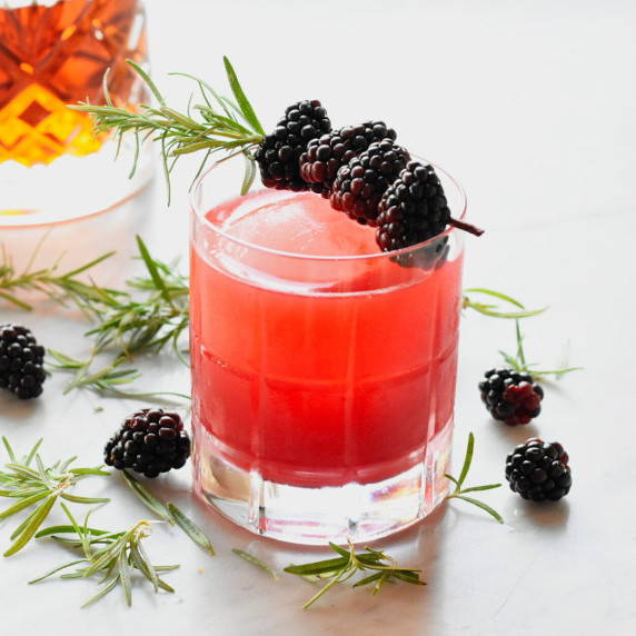 blackberry bour sour in a glass with round ice and blackberries skewered on rosemary
