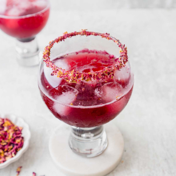 A cocktail glass filled with a blueberry crush mocktail, with a rim of dried rose petals.