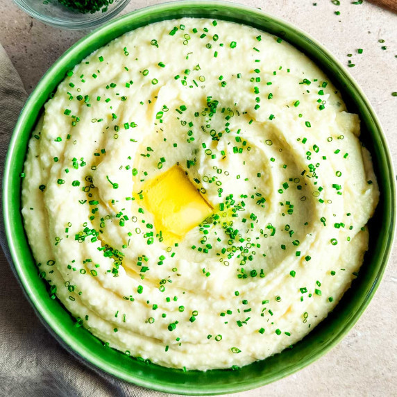 A green ceramic bowl filled with Boursin mashed potatoes topped with a pat of butter and chives.