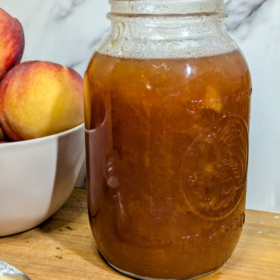 A jar of brandy peach sauce with a bowl of peaches.