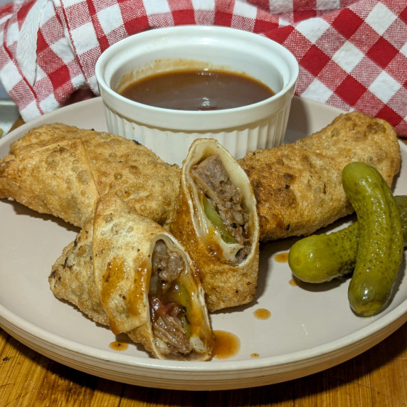 Three brisket egg rolls on a plate. One is cut open in half with BBQ sauce drizzled over it.