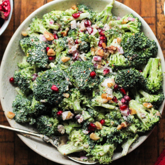 Broccoli pomegranate salad in a white speckled bowl on a brown wooden surface.