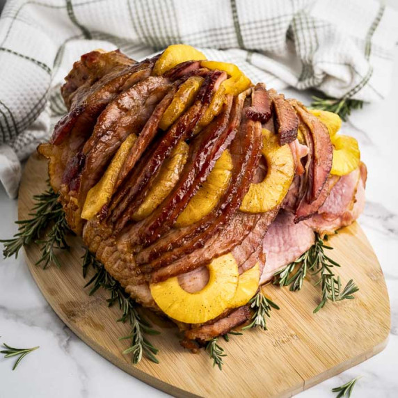 Side shot of ham with pinapple on a wooden cutting board garnished with rosemary sprigs on a marble 