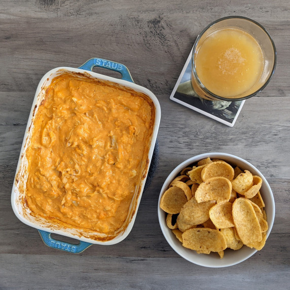 A casserole dish of creamy buffalo chicken dip served with a beer and Fritos scoops