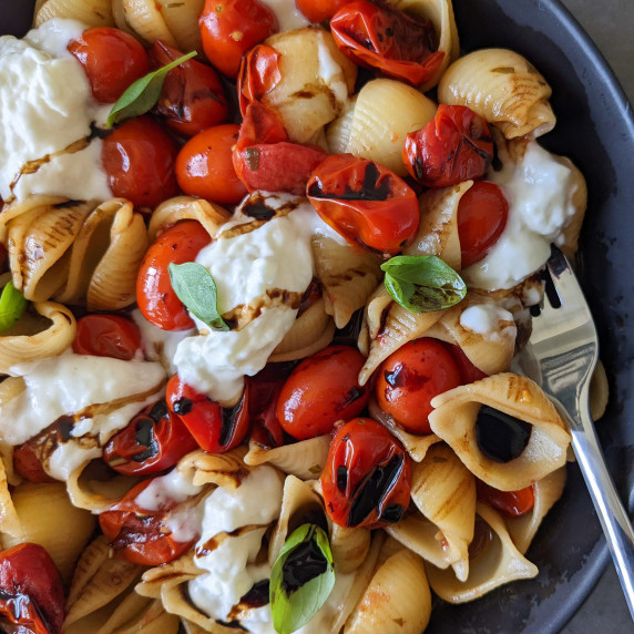 A colorful bowl of pasta shells with blistered tomatoes, burrata cheese, and fresh basil leaves
