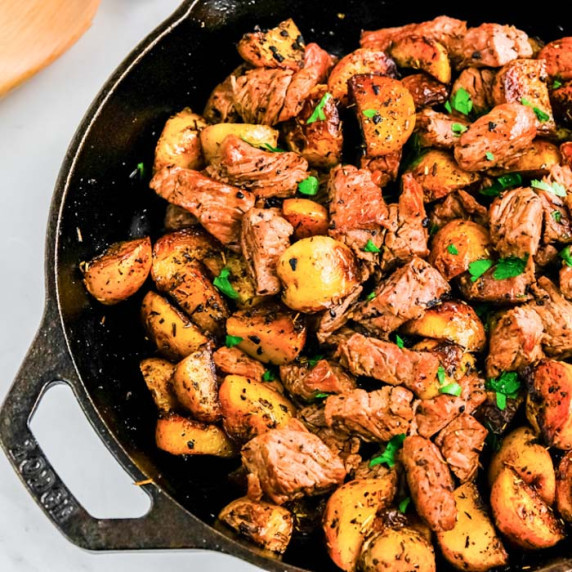 Close up overhead view of steak cubes and potatoes in a black cast iron skillet on a white counter.