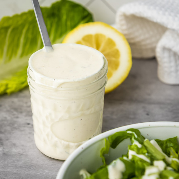 Side shot of caesar dressing in a mason jar with a spoon on the side on a grey surface.