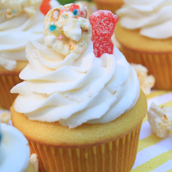 Vanilla cupcakes with Sour Patch Kids candy and Candy Pop on a yellow and white tablecloth.