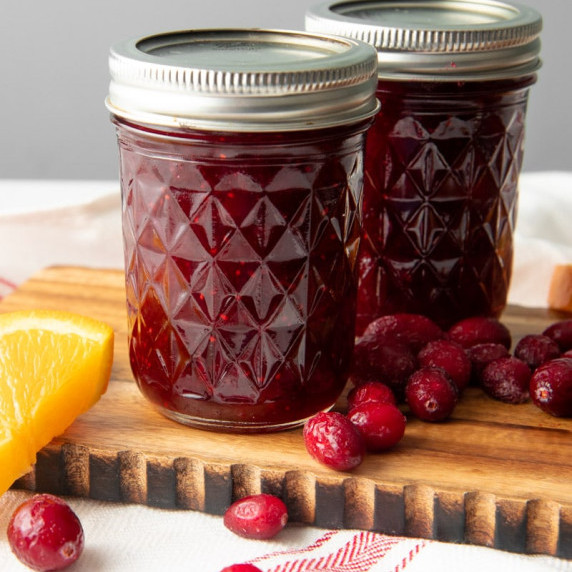 Two quilted canning jars of homemade cranberry sauce stand on a wooden board with fresh cranberries.