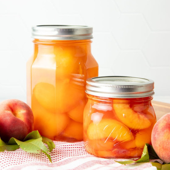 A pint jar of canned peach halves stands next to a half-pint jar of canned peach slices.