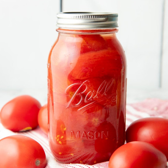 A jar of canned whole tomatoes stands on a red and white kitchen linen surrounded by fresh tomatoes.