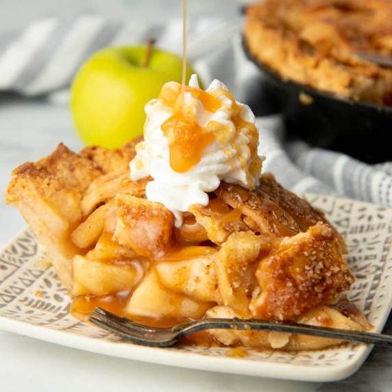 Drizzling extra caramel over a slice of caramel apple pie garnished with whipped cream.