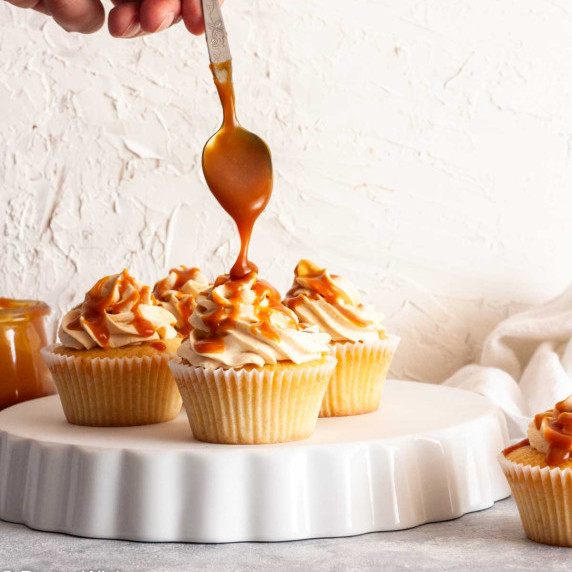 A hand drizzling caramel over caramel filled cupcakes on a white plate.