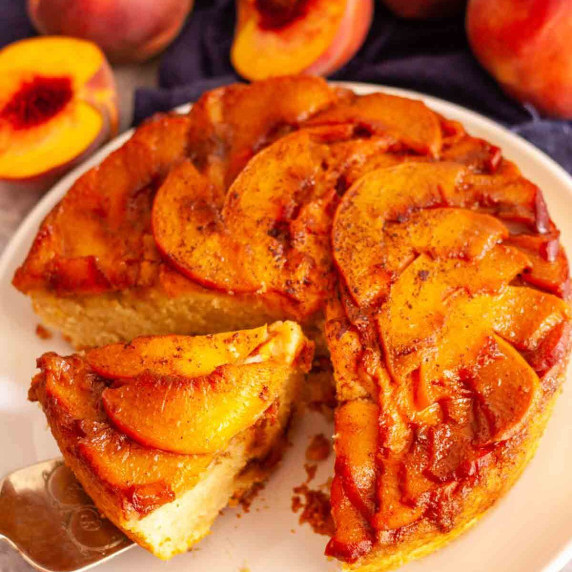 A slice of caramel peach upside down cake on a cake server next to fresh peaches.