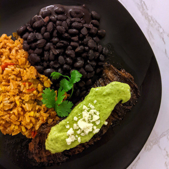 Carne asada steak topped with avocado crema and served with a side of rice and beans