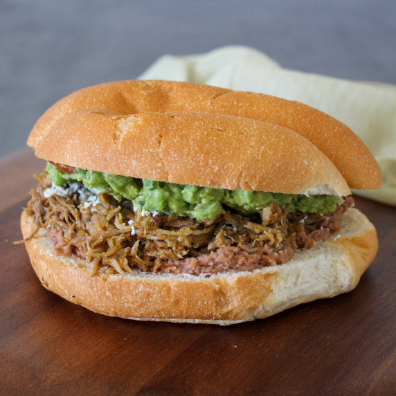 A carnitas torta with refried beans, guacamole, and queso fresco on a fresh telera roll