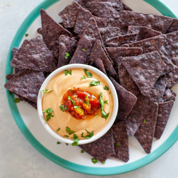 A platter of blue tortilla chips sits with a small bowl of vegan cashew queso nestled into it.