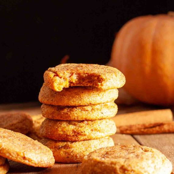 A stack of chewy pumpkin cookies on a wooden surface with a pumpkin and cinnamon sticks in the back.