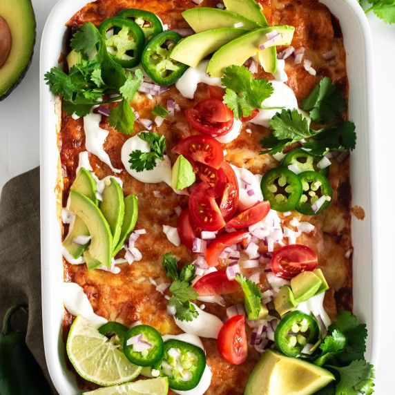 chicken enchiladas in a white baking dish topped with tomatoes, onion, avocado and sour cream.