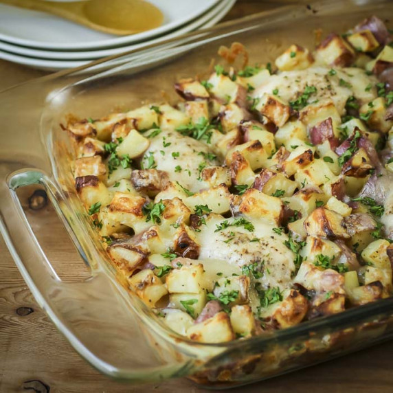 Side shot of chicken potato bake in a casserole dish on a wooden table with plates in the background