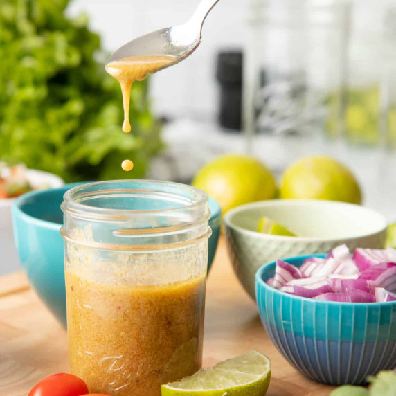 A spoon scoops up chili lime vinaigrette from a mason jar surrounded by bowls of fresh ingredients.