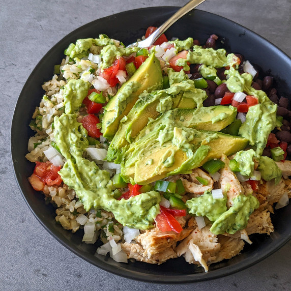 A burrito bowl of rice, black beans, chicken, and fresh produce topped with avocado slices