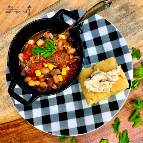 A small cast iron pot filled with vegan chili on a checkered plate with a slice of cornbread.