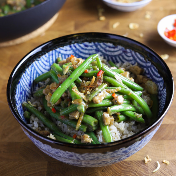 photo of a bowl of chinese green beans and tempeh (vegan/gf) on a wooden table with a pan behind.