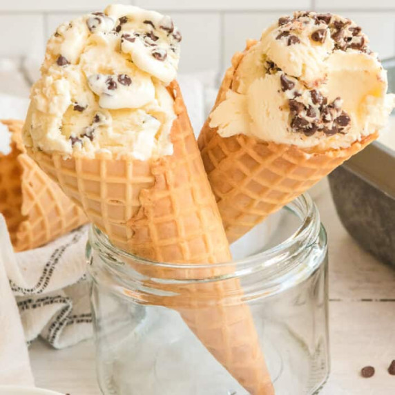 Two waffle cones resting in a glass container full of chocolate chip ice cream, view from the side.