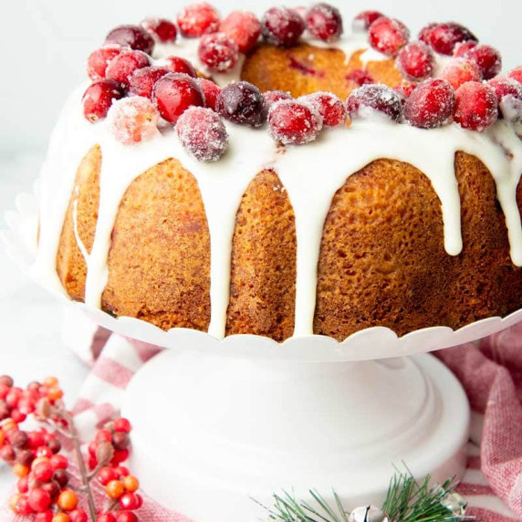 A Christmas bundt cake topped with sugared cranberries and a white glaze sits on a white cake stand.