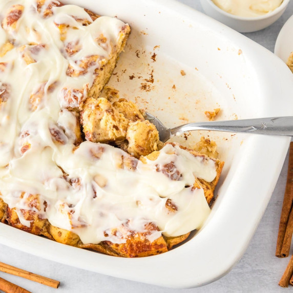 A white dish with frosted cinnamon roll casserole, partially served as a spoon scoops from the pan.