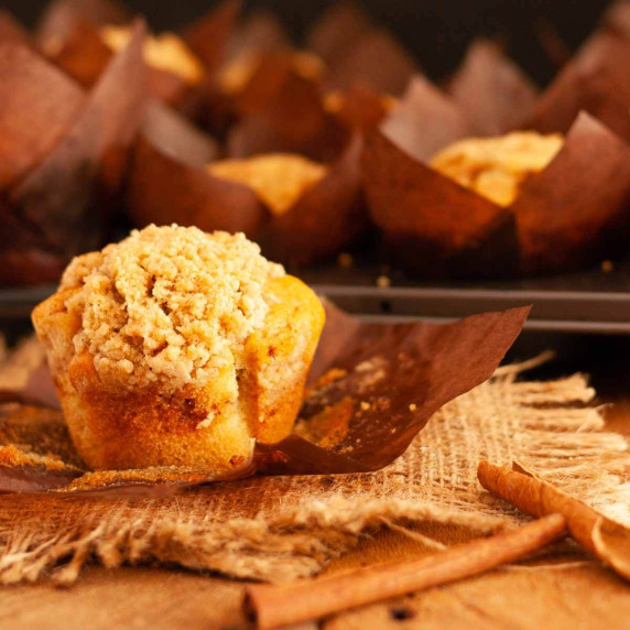 A cinnamon streusel muffin on burlap on a wooden surface next to cinnamon sticks and muffins.