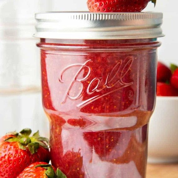 Close-up of homemade strawberry jam in a stacking mason jar with fresh strawberries beside it.