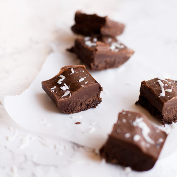 Small squares of vegan chocolate fudge sprinkled with coconut sit on a white countertop.